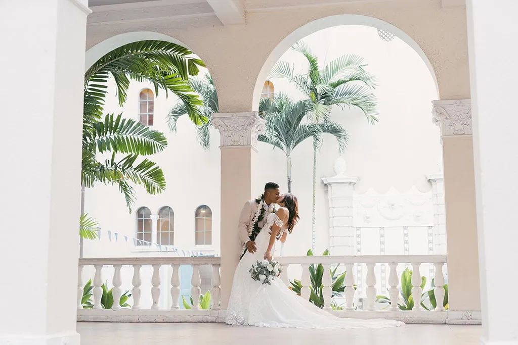 A bride and groom kissing near a large archway window with coconut trees in the background, captured by Rachel Kathryn Photo